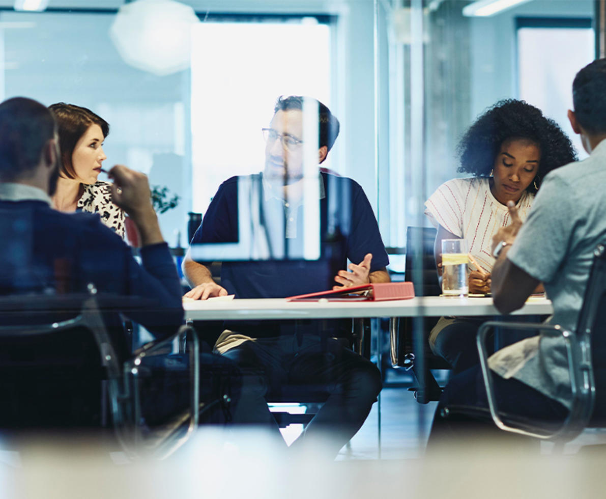 view through glass of 5 people sitting at a conference table working